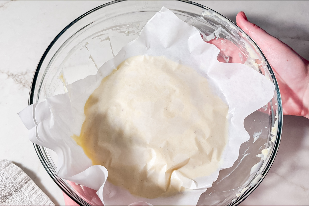 A glass bowl lined with parchment paper and filled with a creamy, light-colored pastry cream is held by two hands over a white surface.