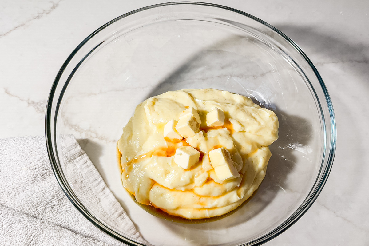 A glass bowl containing pastry cream topped with cubes of butter and vanilla extract, placed on a white countertop next to a white towel.