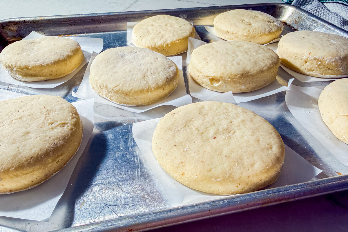 Unbaked biscuit dough rounds rest on pieces of parchment paper on a metal baking sheet, ready to be baked. Sunlight highlights the texture and surface of the dough.