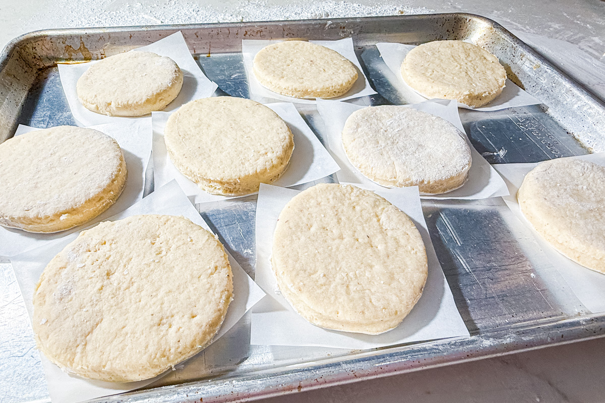 Unbaked biscuit dough rounds spaced on parchment squares, arranged on a metal baking sheet, ready to go into the oven.