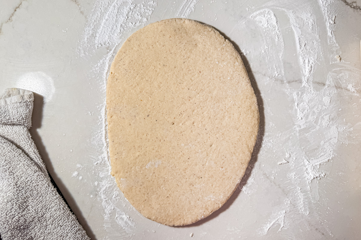 An oval piece of dough is rolled out on a floured surface with a textured cloth partially visible on the left side.