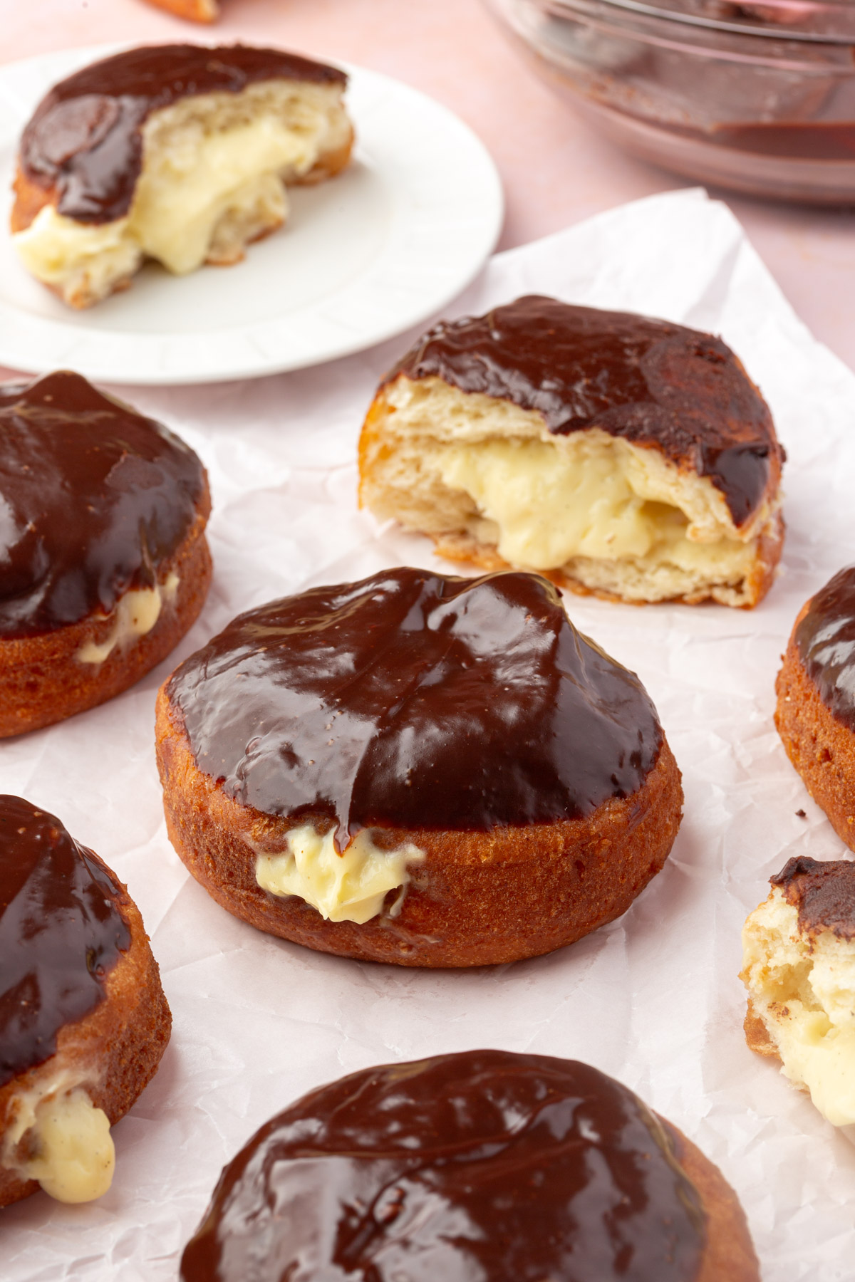 Several chocolate-glazed cream-filled doughnuts sit on parchment paper, with one doughnut cut open to show the creamy vanilla filling inside. A plate with a half-eaten doughnut is in the background.