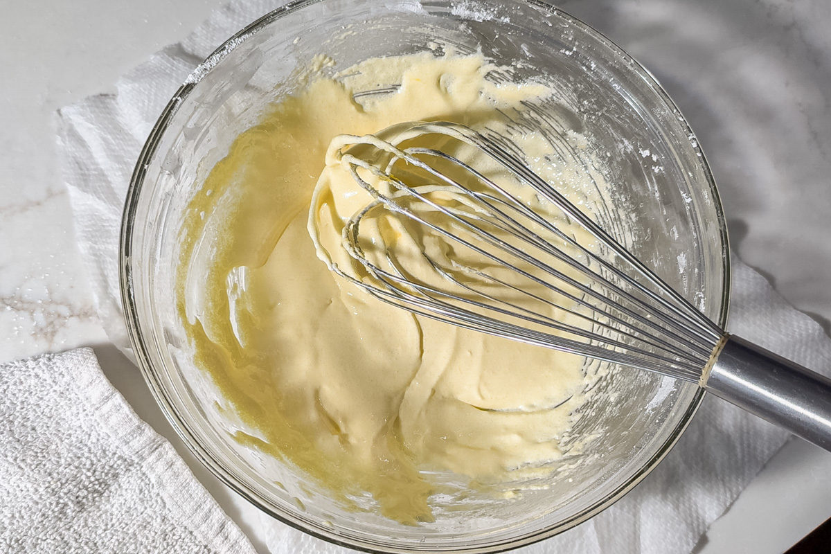 A glass bowl containing pale yellow whisked egg yolks being mixed with a metal whisk, placed on a white cloth on a marble surface.