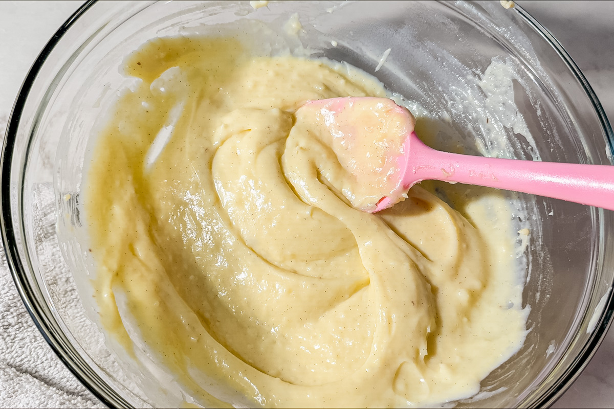 A glass bowl filled with creamy, pale yellow pastry cream being mixed with a pink silicone spatula, resting on a white surface.