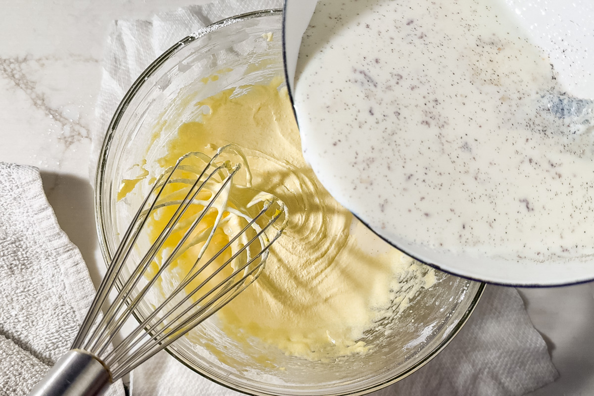 A metal whisk rests in a glass bowl of yellow egg yolk mixture while a pot pours whole milk with vanilla specks into the bowl, set on a white surface with a textured cloth nearby.