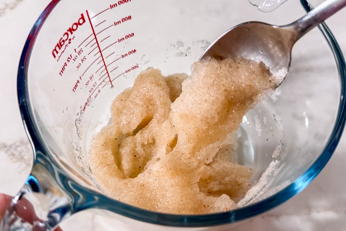 A close-up of a glass measuring cup containing a thick, beige, gelatinous mixture being stirred with a metal spoon. The cup has red measurement markings on the side.