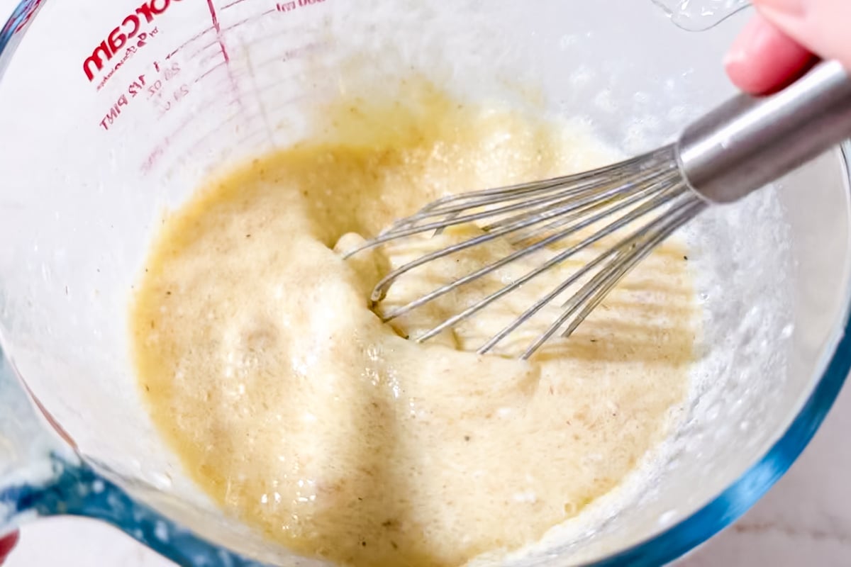 A close-up of a person whisking a frothy, yellow batter in a glass measuring cup. The mixture appears to be in the process of being blended for baking or cooking.