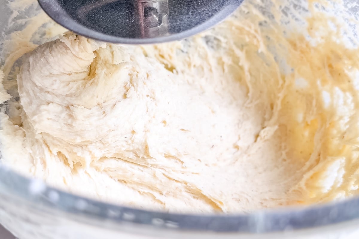 Close-up of a stand mixer blending thick, creamy dough in a glass mixing bowl. The dough appears soft and partially mixed, with the metal paddle attachment visible at the top.