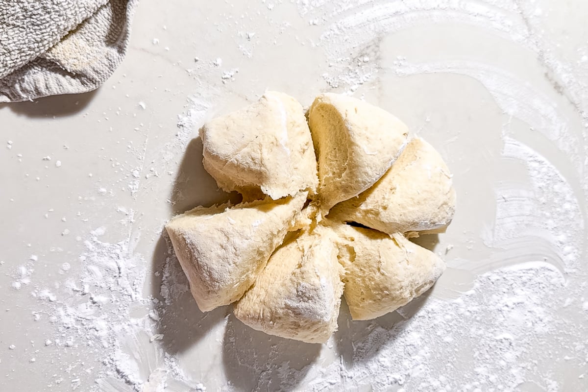 A ball of biscuit dough divided into eight wedges sits on a floured surface, with a kitchen towel partially visible in the corner.