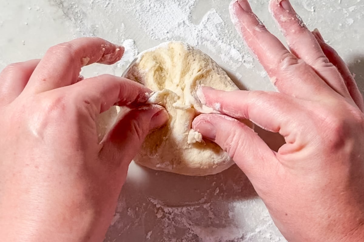 Two hands knead a small ball of dough on a floured surface, pinching and folding the soft dough while flour is scattered around.