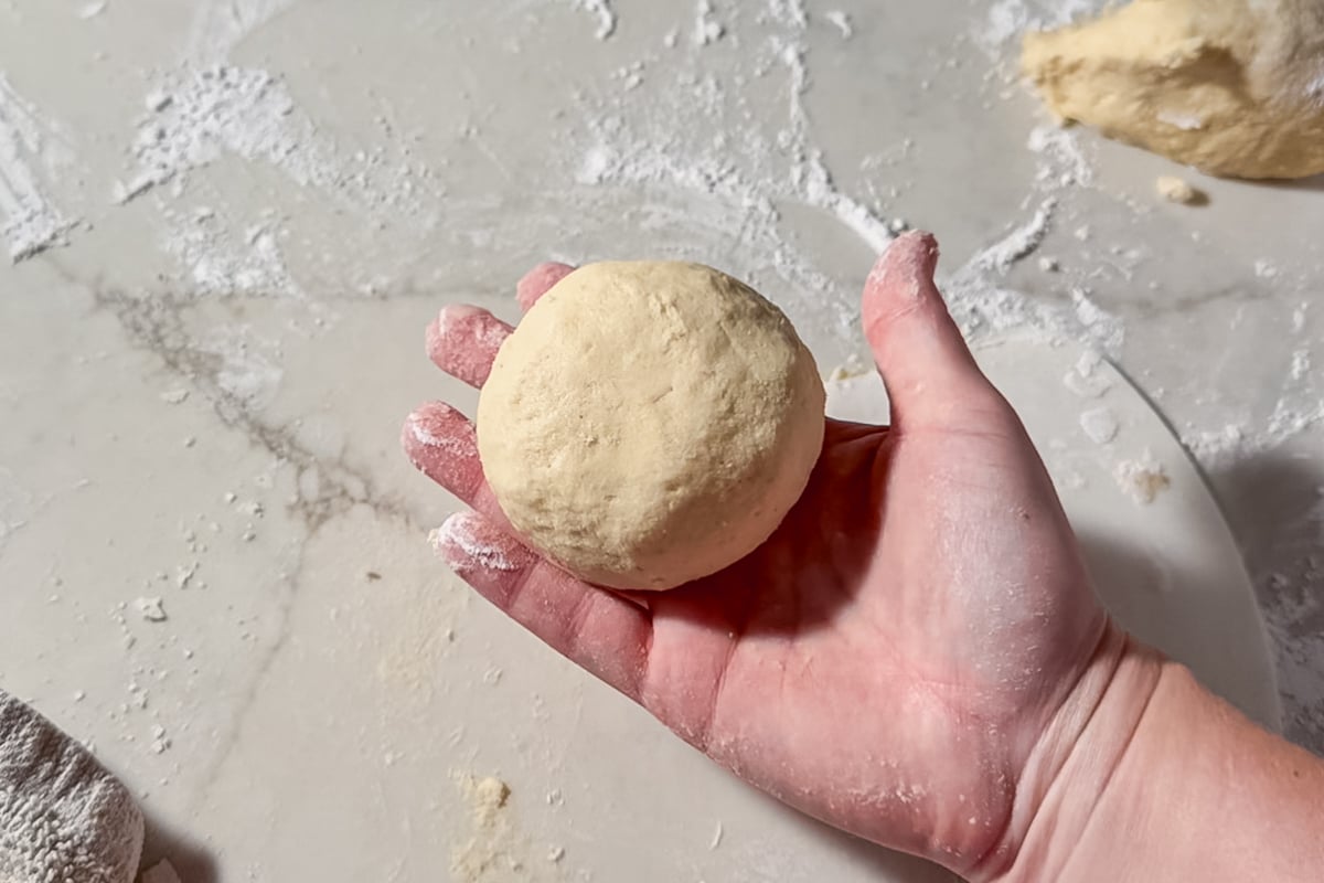 A hand holding a small, round ball of dough over a floured surface, with more dough and flour visible in the background.
