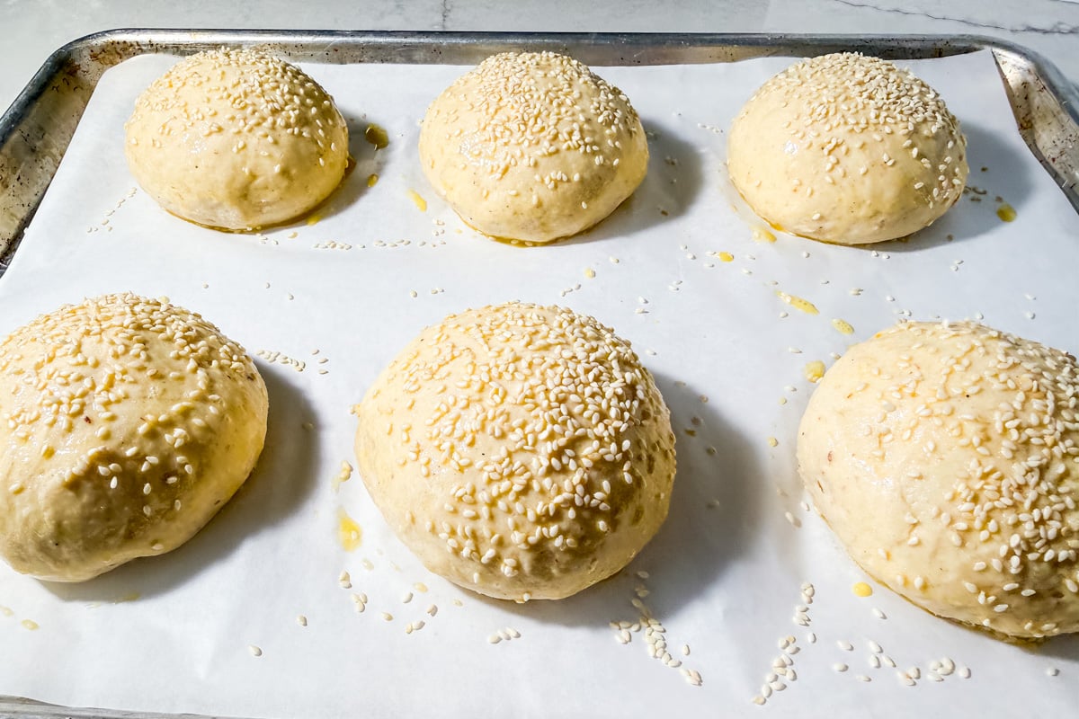 Six unbaked, round dough buns topped with sesame seeds are arranged on a parchment-lined baking sheet, ready to be baked.