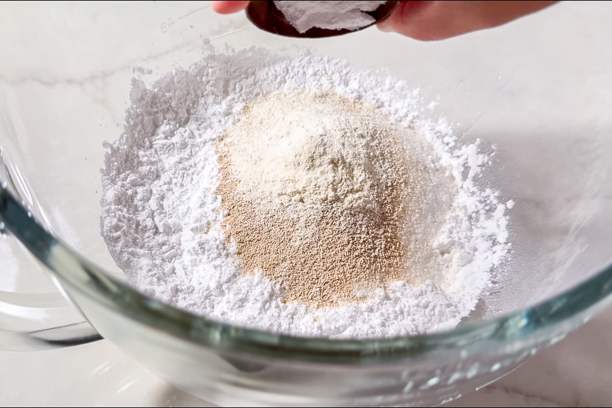 A glass mixing bowl filled with white flour and yeast, with a hand holding a spoonful of baking powder above the bowl on a white countertop.