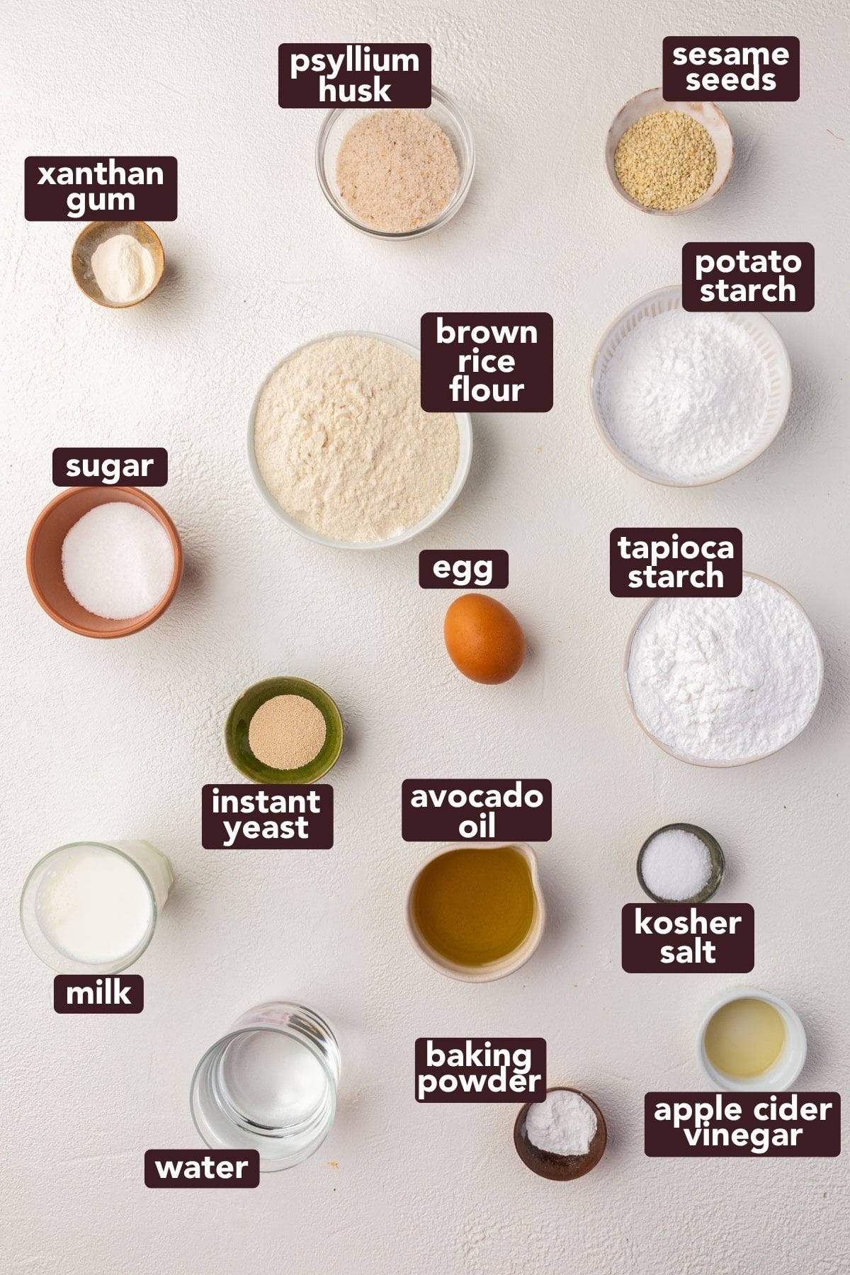 Various labeled baking ingredients including brown rice flour, potato starch, tapioca starch, egg, milk, water, instant yeast, avocado oil, baking powder, apple cider vinegar, kosher salt, sugar, xanthan gum, psyllium husk, and sesame seeds.