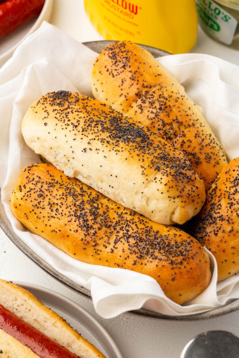 A basket lined with a white cloth holds several poppy seed-topped gluten-free hot dog buns. Other food items, including part of a hot dog and condiment bottles, are visible in the background.