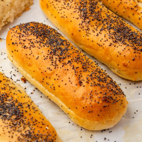 Four golden brown gluten-free hot dog buns topped with poppy seeds are arranged on a white parchment paper surface.
