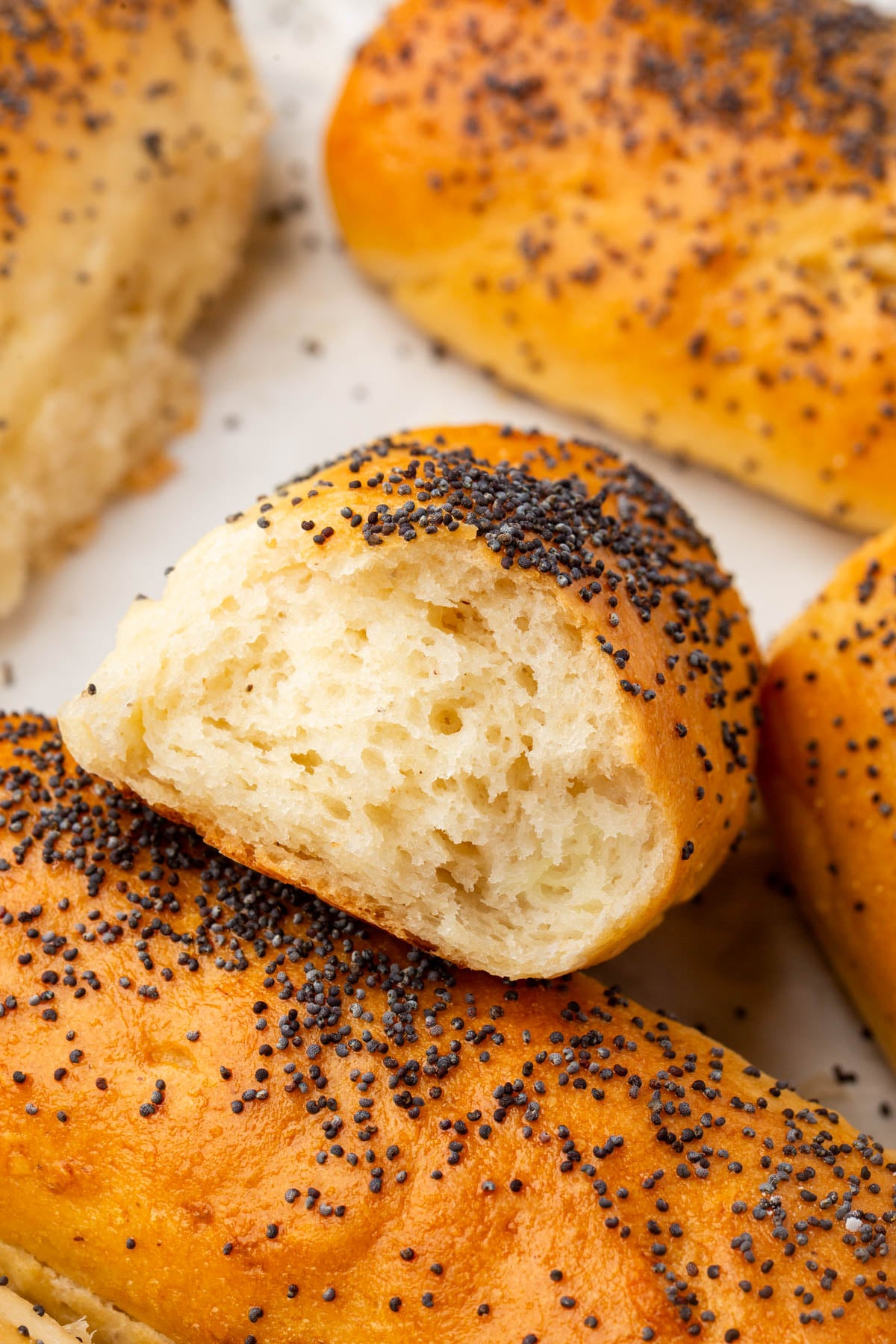 A close-up of soft, golden brown gluten-free hot dog rolls topped with poppy seeds, one of which is broken open to show the fluffy, airy inside.