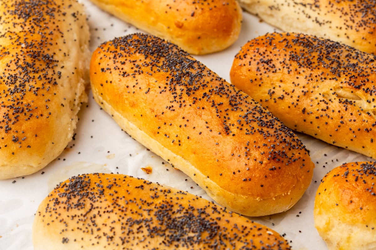 Golden brown gluten-free hot dog buns topped with poppy seeds are arranged closely together on a sheet of parchment paper. The rolls have a soft texture and a slightly glossy, baked surface.