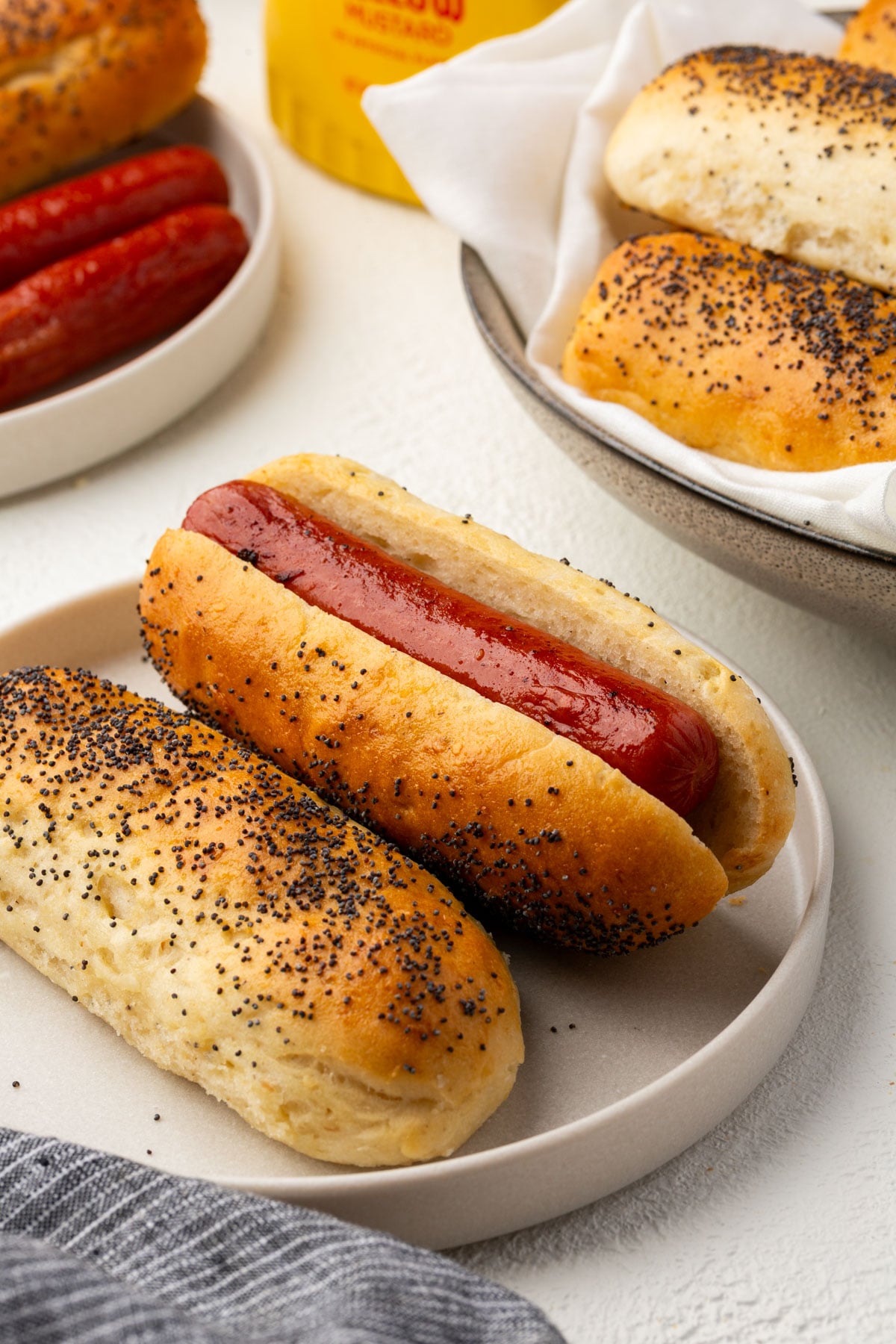 Two hot dogs in poppy seed buns on a plate, with another plate of poppy seed buns and a bottle of mustard in the background. A striped cloth is partially visible in the corner.