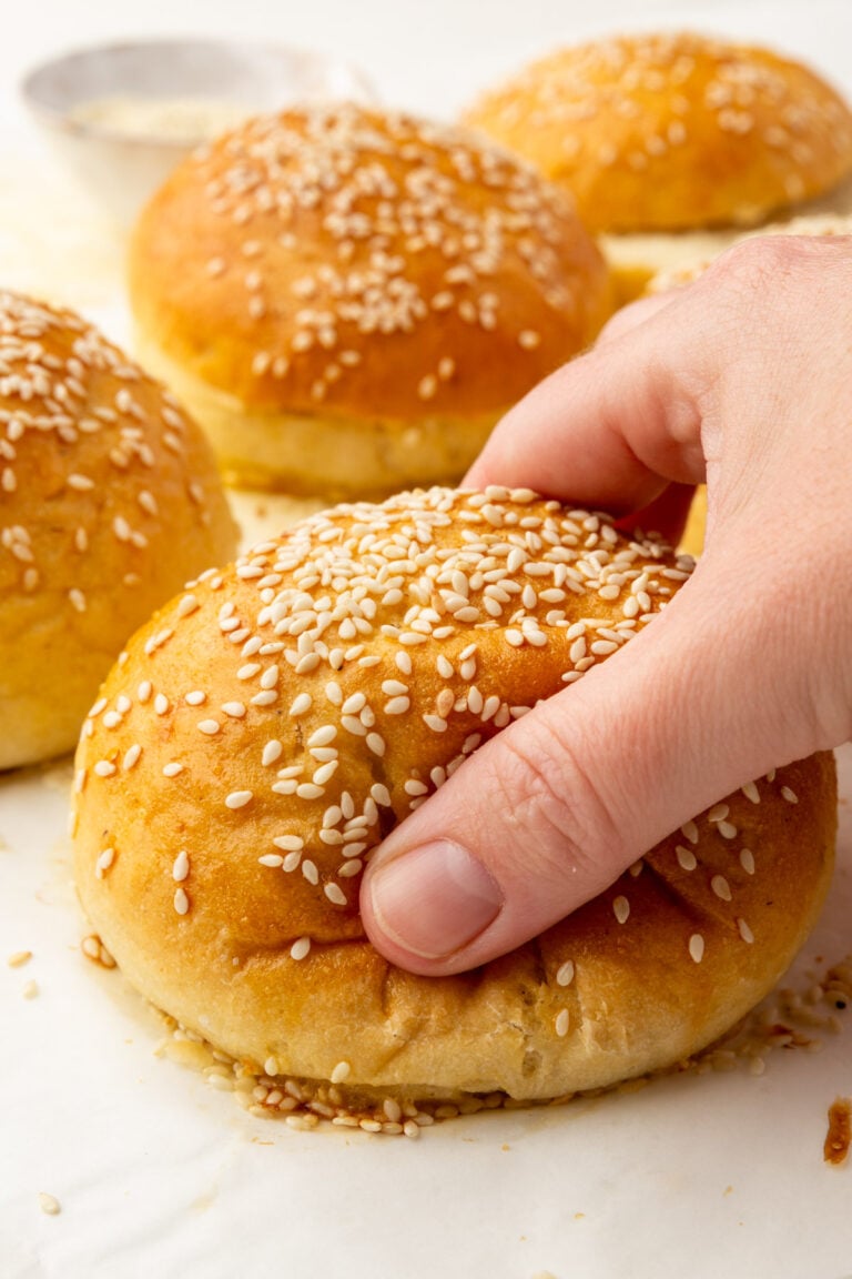 A hand picking up a round, golden-brown gluten-free sesame seed hamburger bun from a group of buns on a white surface.