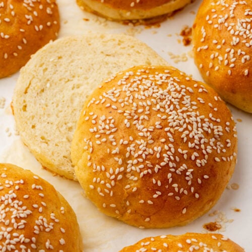 Golden brown gluten-free sesame seed hamburger buns are arranged on a white surface, with one bun sliced open to show the soft, airy interior.