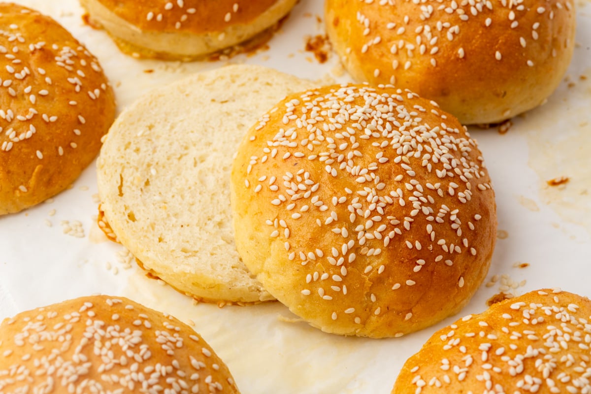 Several golden brown gluten-free hamburger buns with sesame seeds on a white surface, with one bun cut in half to show its soft interior.
