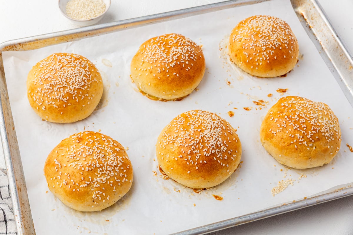 A baking tray lined with parchment paper holds six golden brown, round gluten-free hamburger buns topped with sesame seeds. A small dish of sesame seeds is visible in the background.