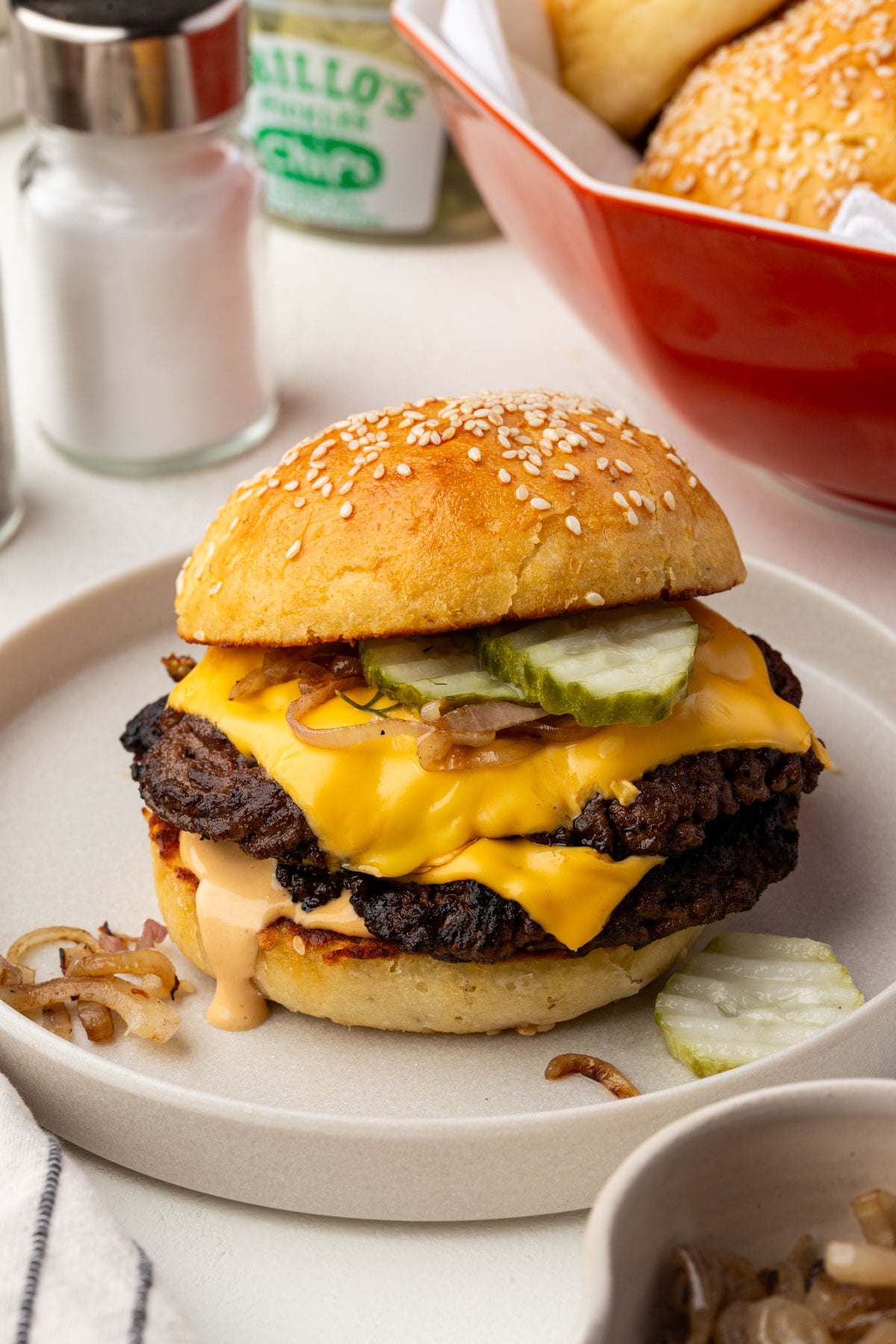 A gluten-free cheeseburger with two beef patties, melted cheese, pickles, grilled onions, and sauce on a gluten-free sesame seed bun sits on a plate. A salt shaker and a dish of buns are in the background.