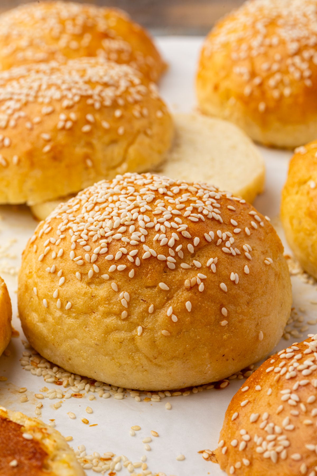 A close-up of golden-brown gluten-free hamburger buns topped with sesame seeds, arranged on a sheet of parchment paper. One bun in the background is sliced open to show its soft, airy interior.