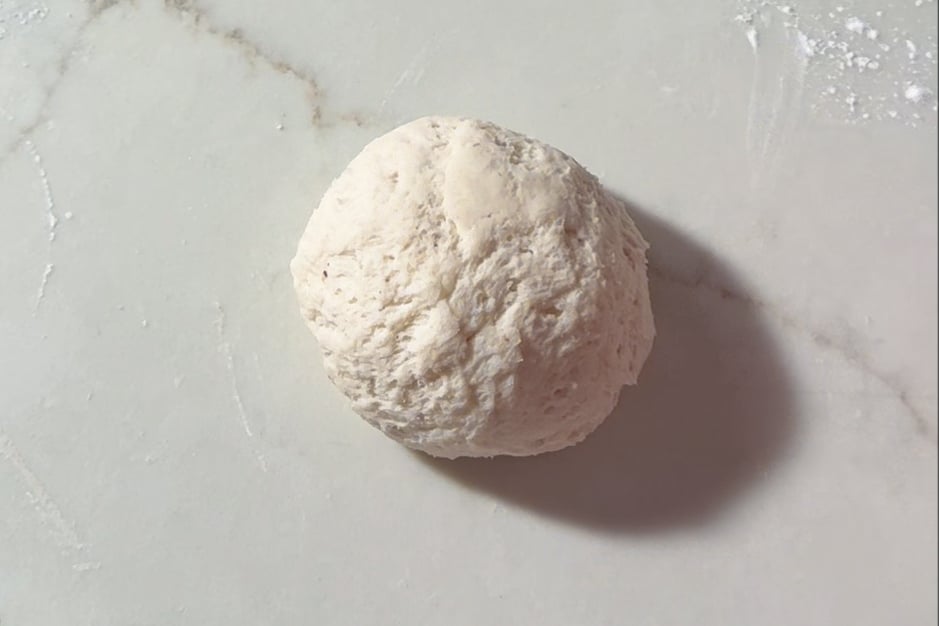 A ball of raw bagel dough sits on a lightly floured, white marble surface, casting a soft shadow to the right.