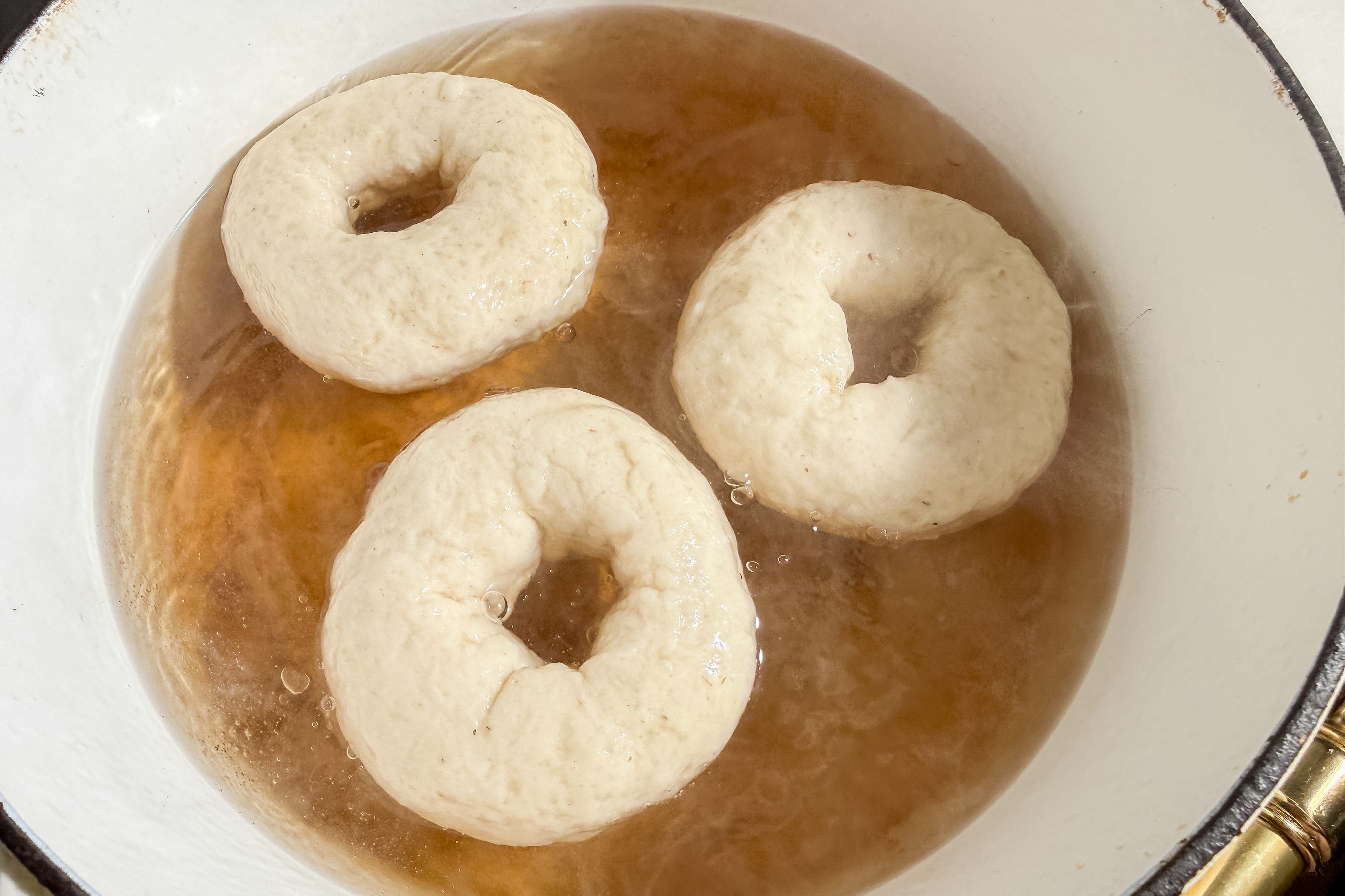 Three proofed bagels are boiling in a pot of water mixed with brown sugar, shown from above.