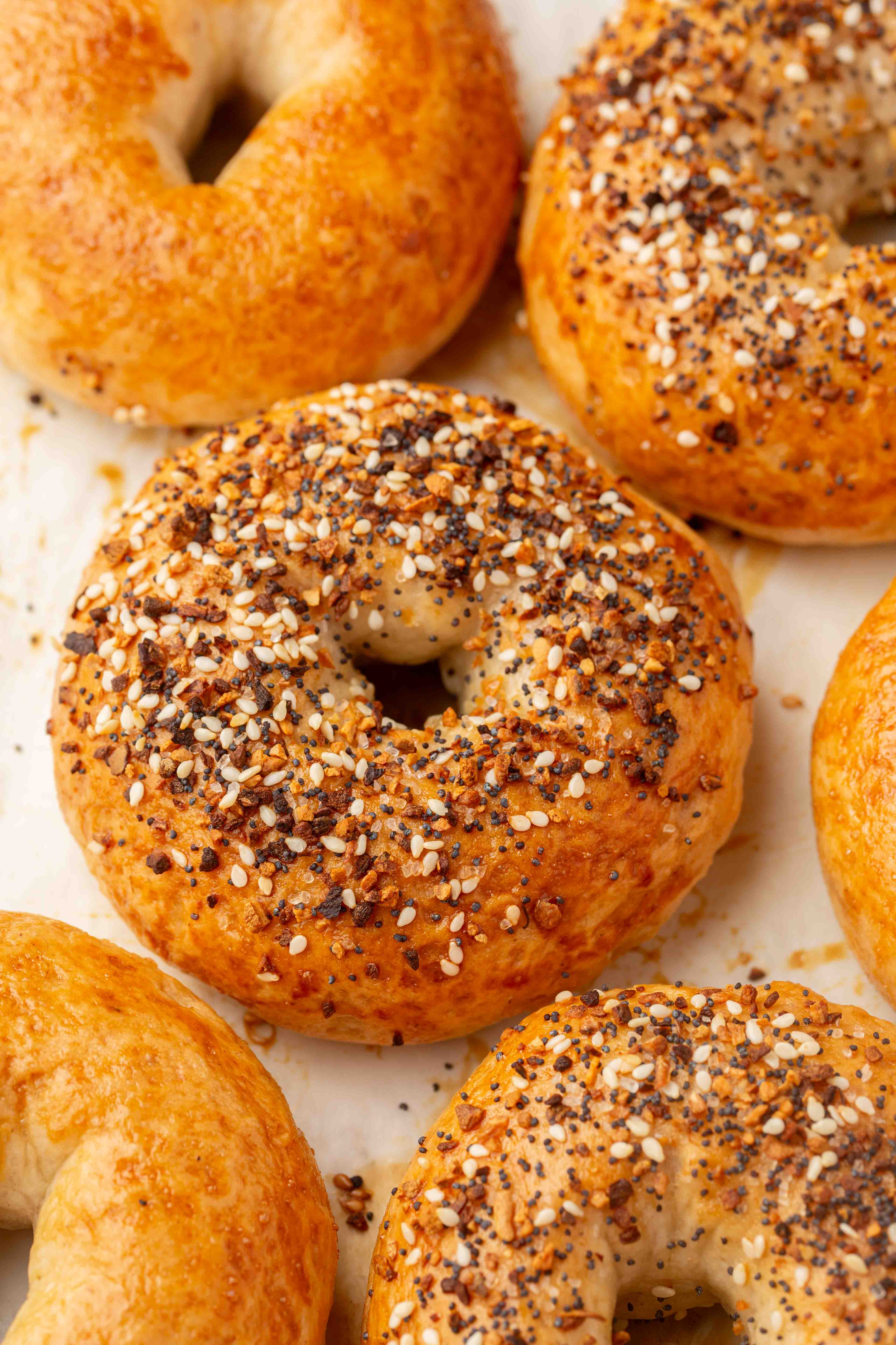 Golden gluten-free bagels, some topped with sesame seeds and seasoning, rest on a white surface. The bagels have a glossy finish and are freshly baked.