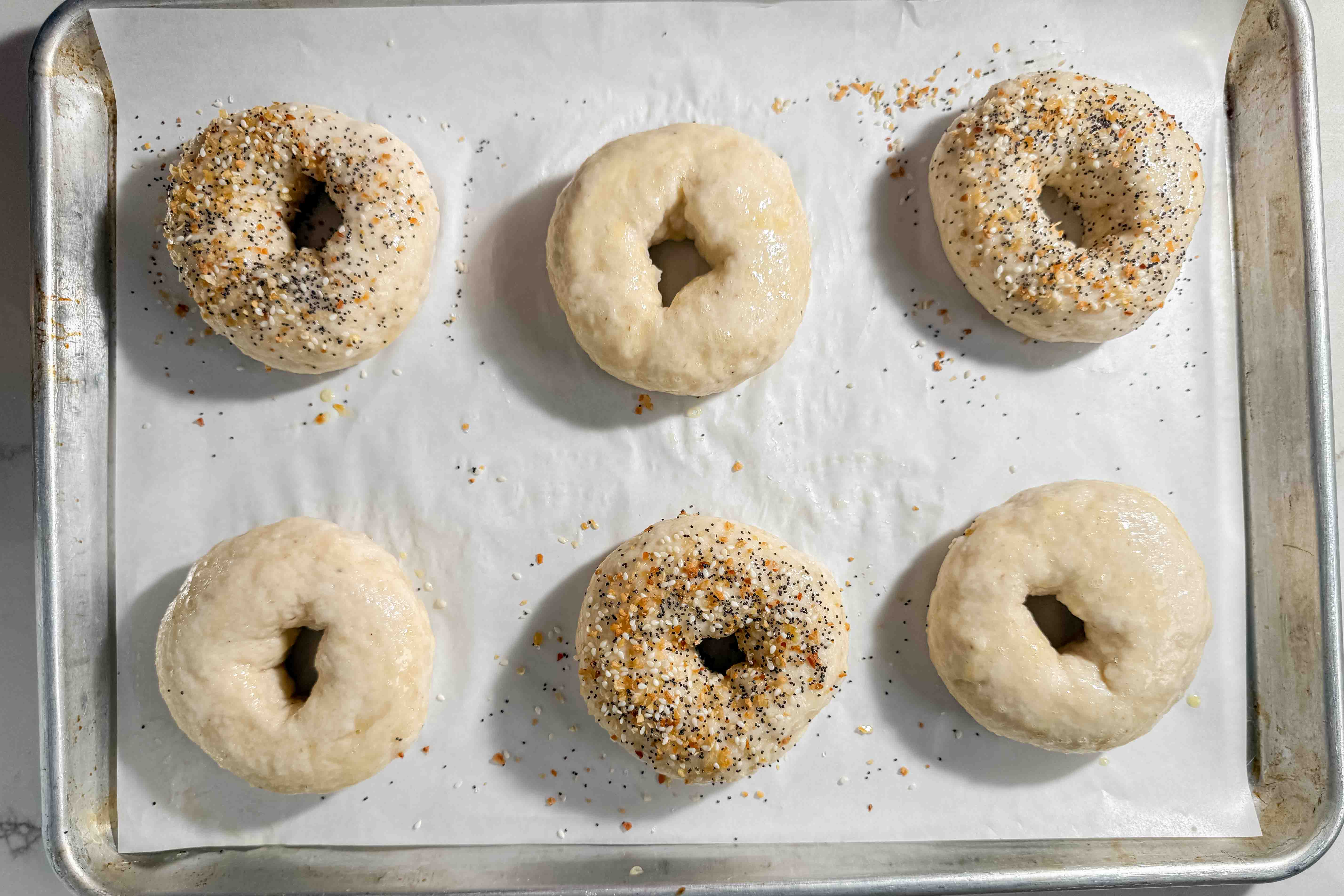 Six unbaked bagels on a parchment-lined baking sheet, three topped with seasoning and three plain, arranged in two rows of three.