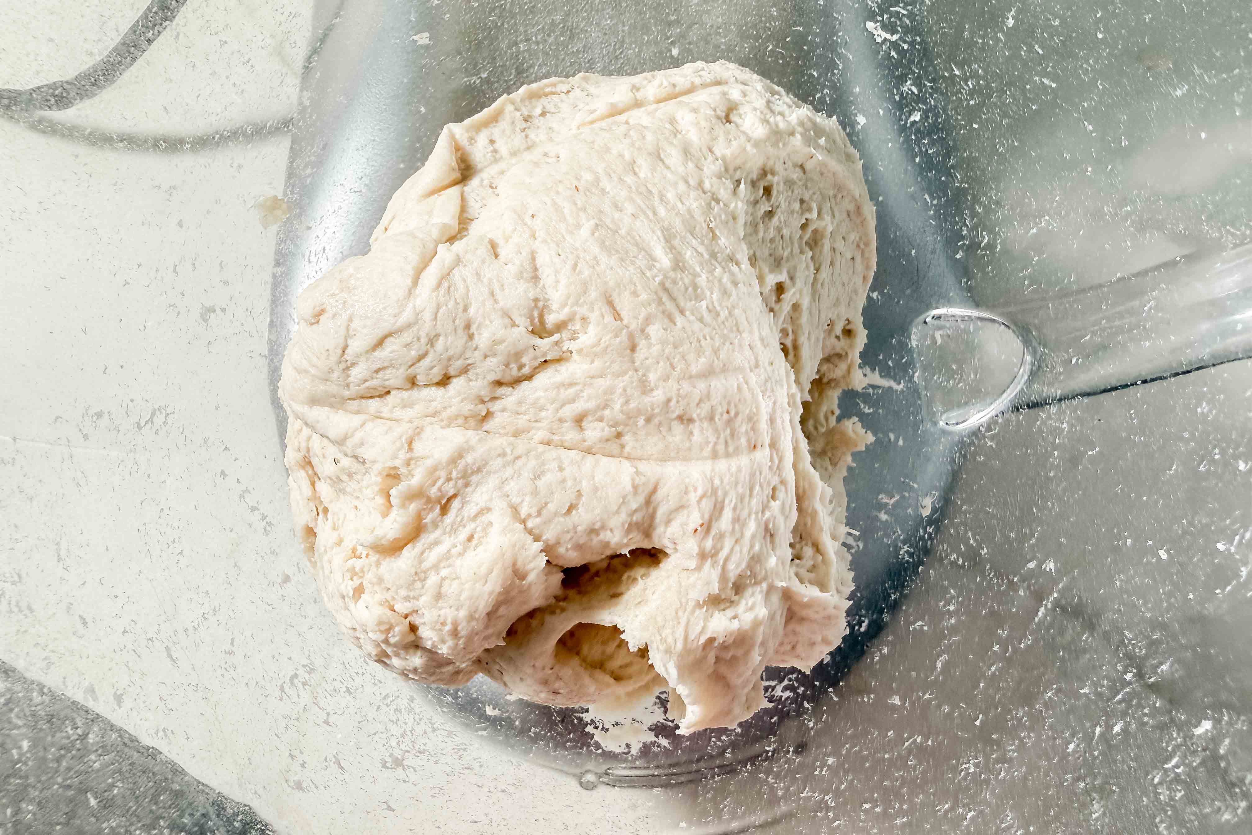 A ball of gluten-free bagel dough sits in a glass mixing bowl. The dough is soft and sticky, and some gluten-free flour is visible on the sides of the bowl.