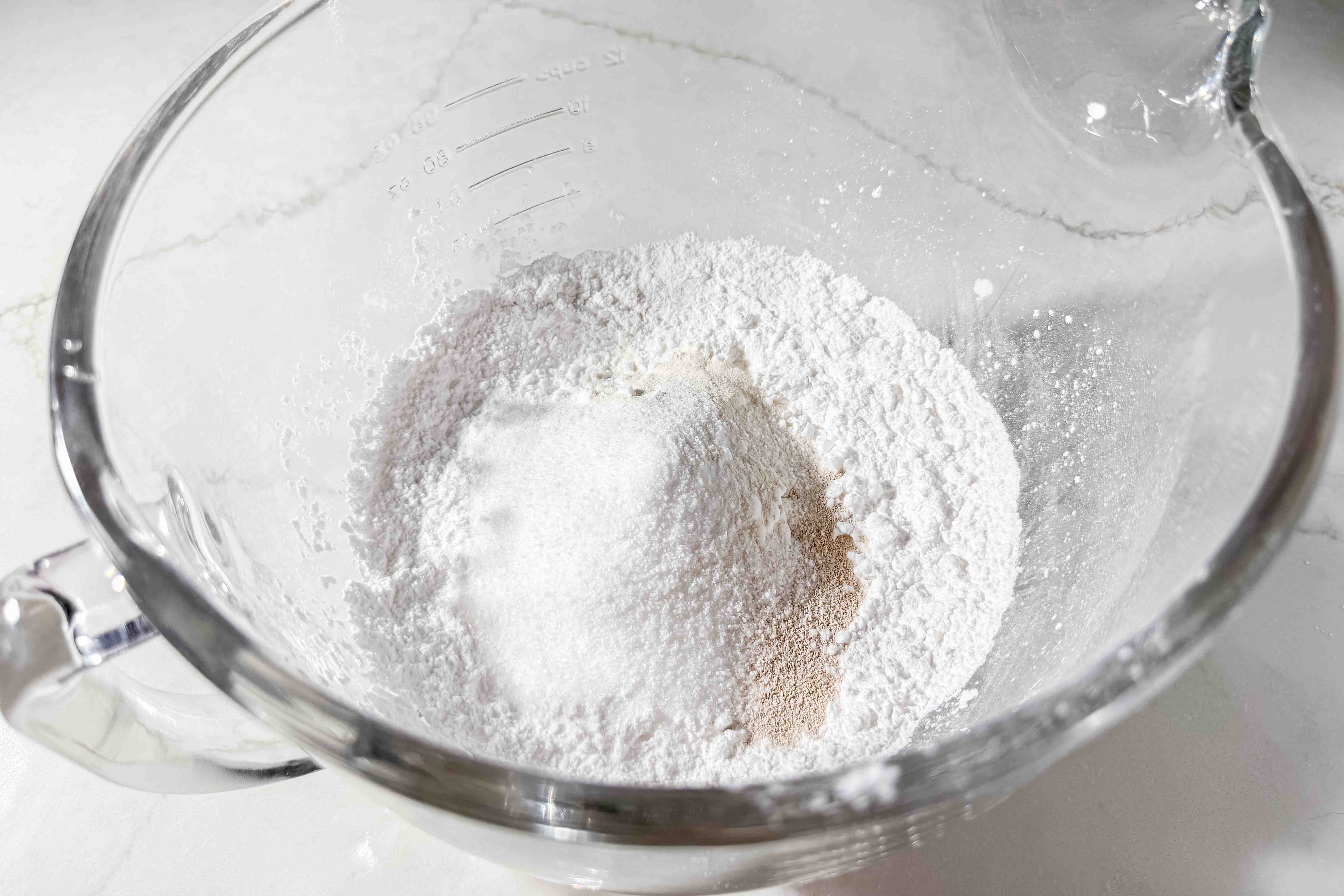 A clear glass mixing bowl containing a mound of gluten-free flour, sugar, and yeast on a white countertop, ready to be mixed for baking.