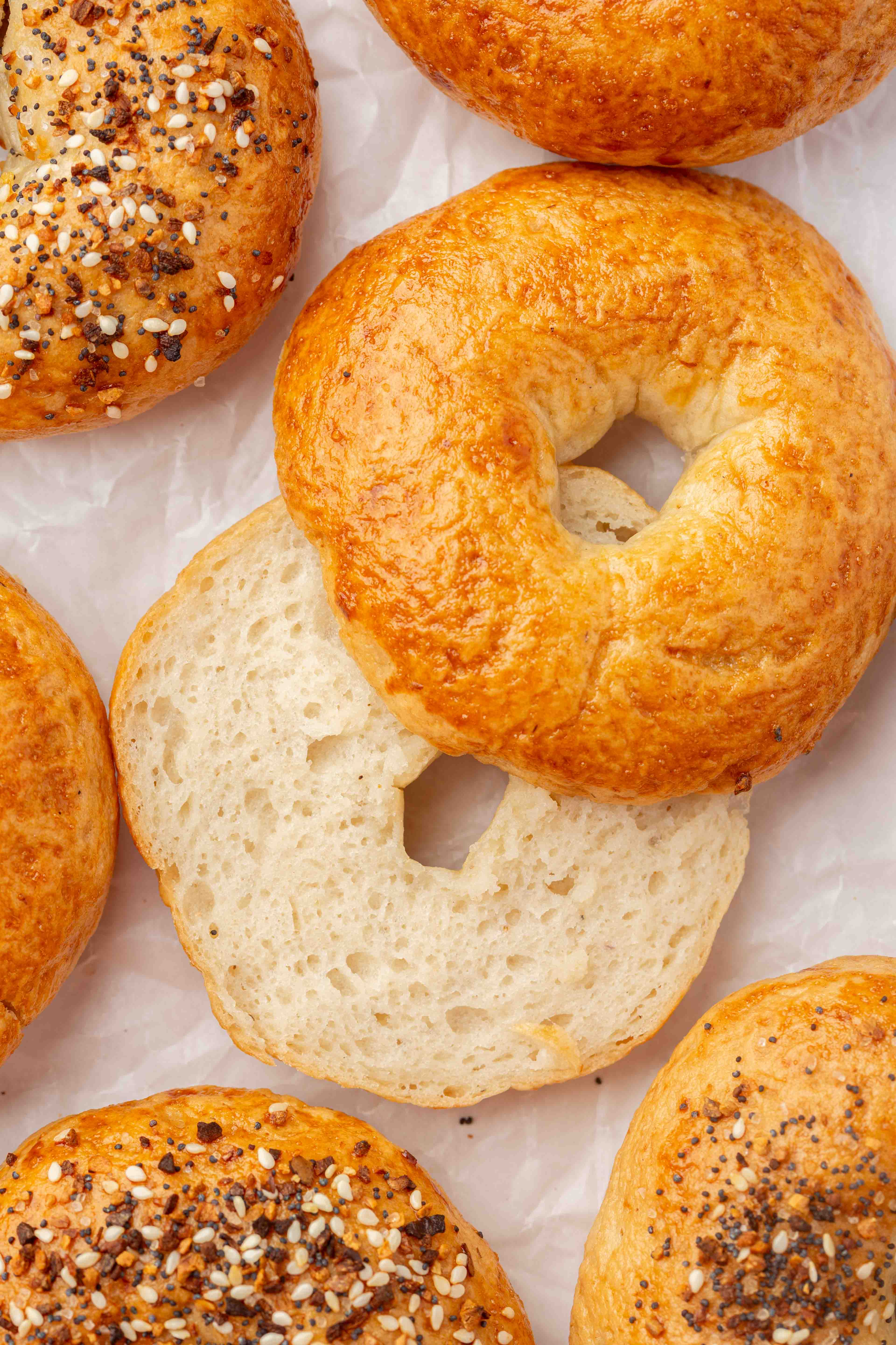 Several golden brown gf bagels, some with everything seasoning and one plain bagel sliced open, are arranged on crumpled white parchment paper.