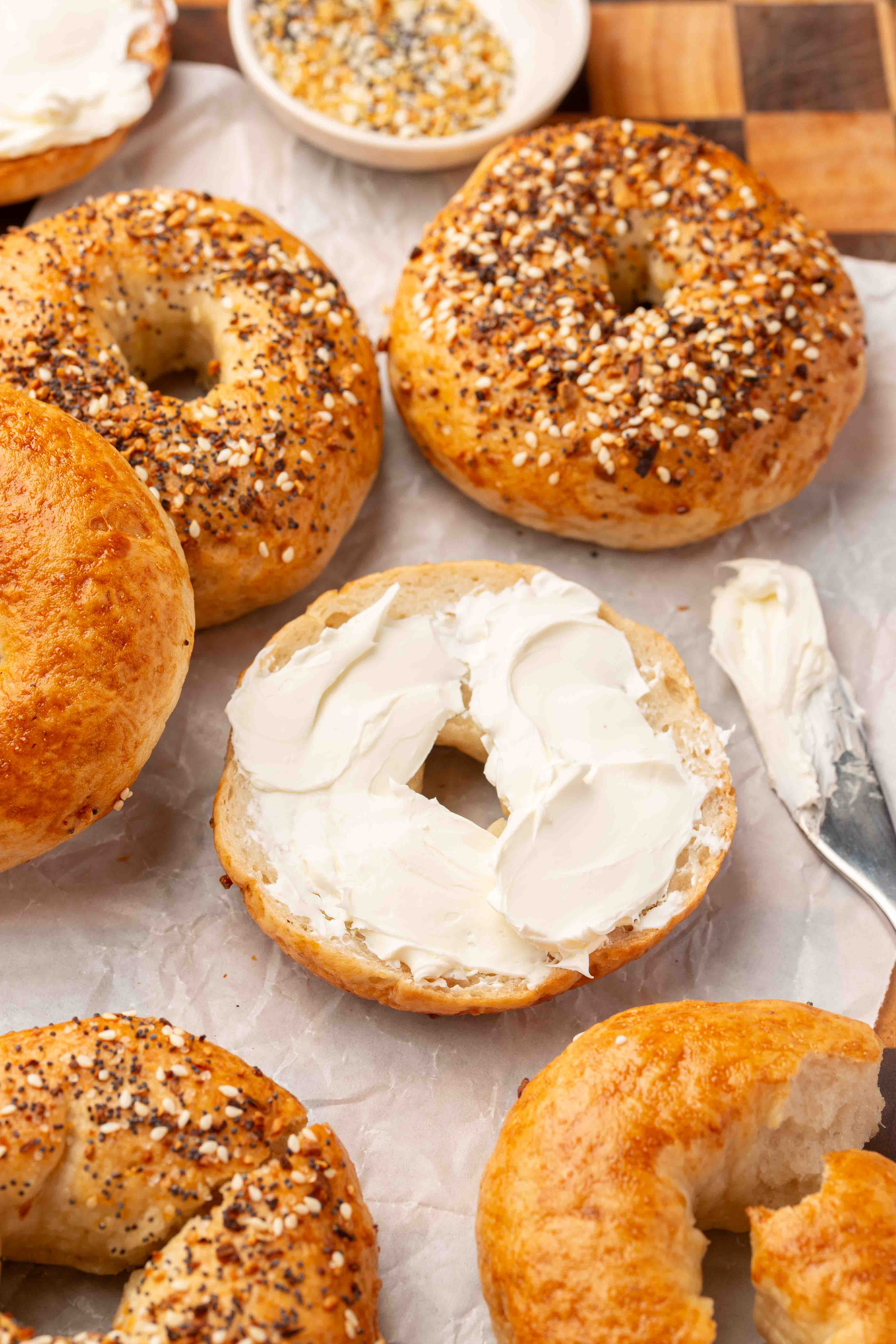 A sliced bagel spread with cream cheese sits among whole plain and everything gluten-free bagels on parchment; a knife with cream cheese rests nearby on a wooden surface.