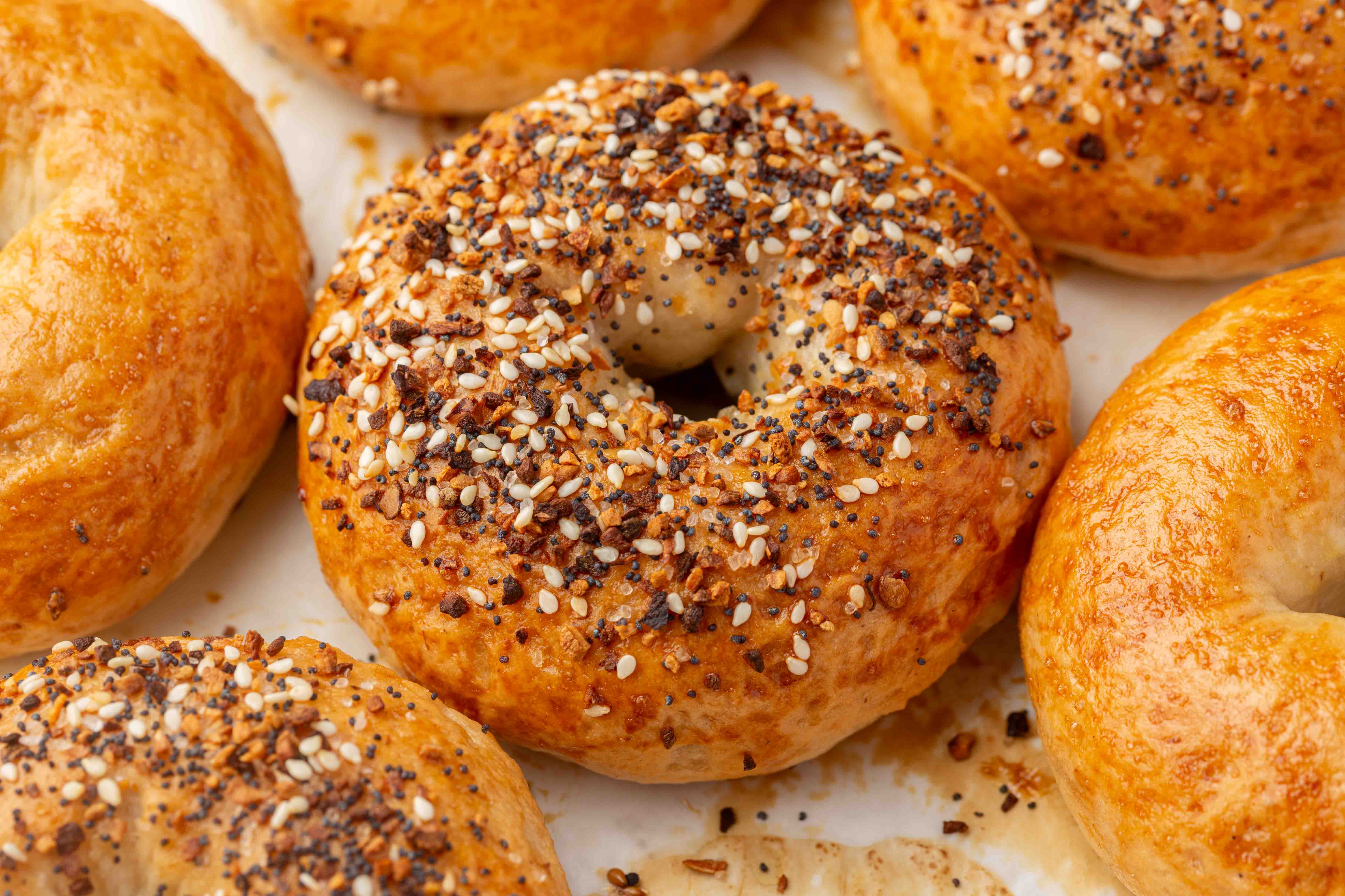 A close-up of golden brown gluten-free bagels, some topped with a mix of sesame and poppy seeds, arranged on a parchment-lined surface.