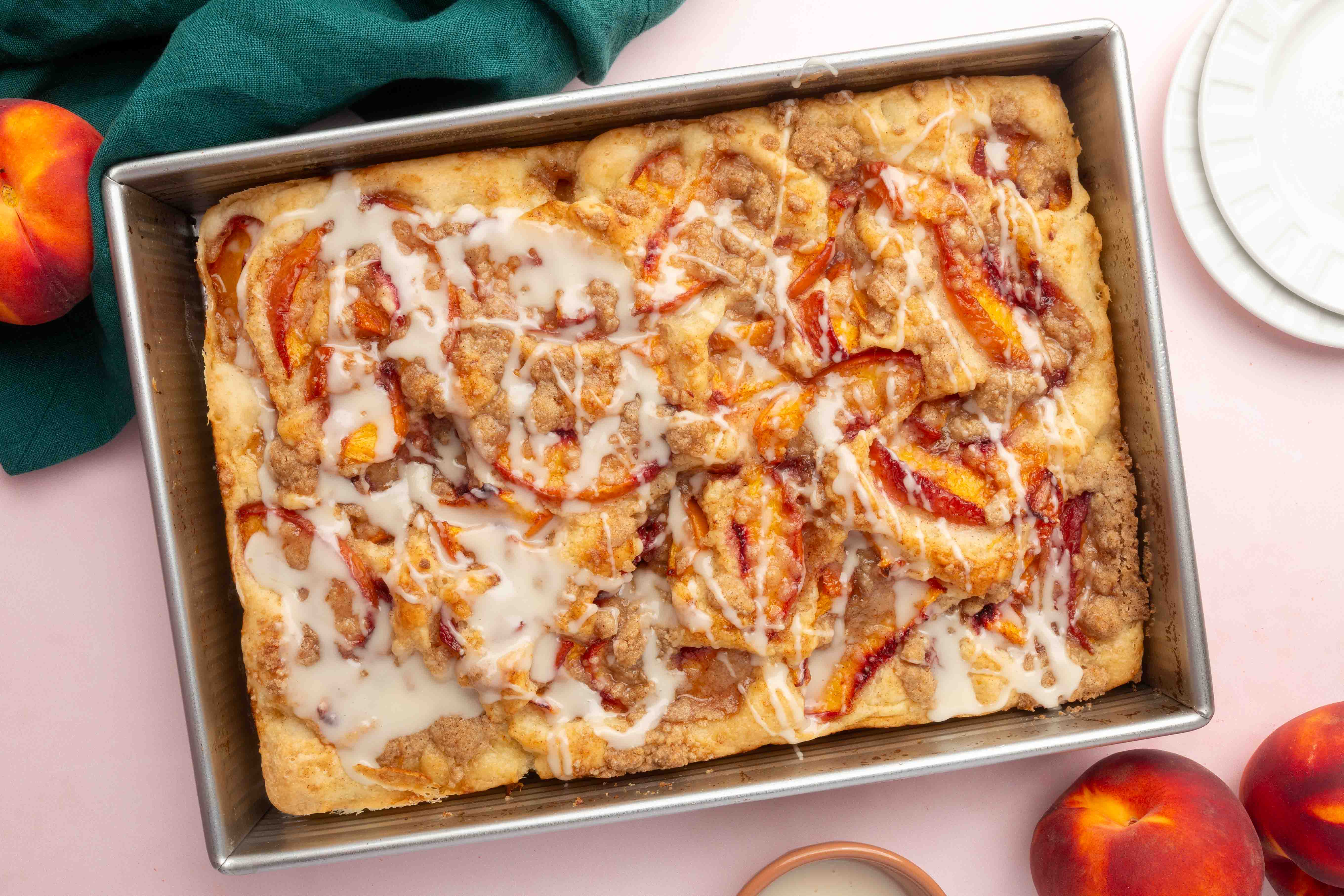 Rectangular pan with Gluten-Free Peach Crumble Focaccia topped with icing and crumble, surrounded by fresh peaches, a green cloth, and a white plate on a pink surface.