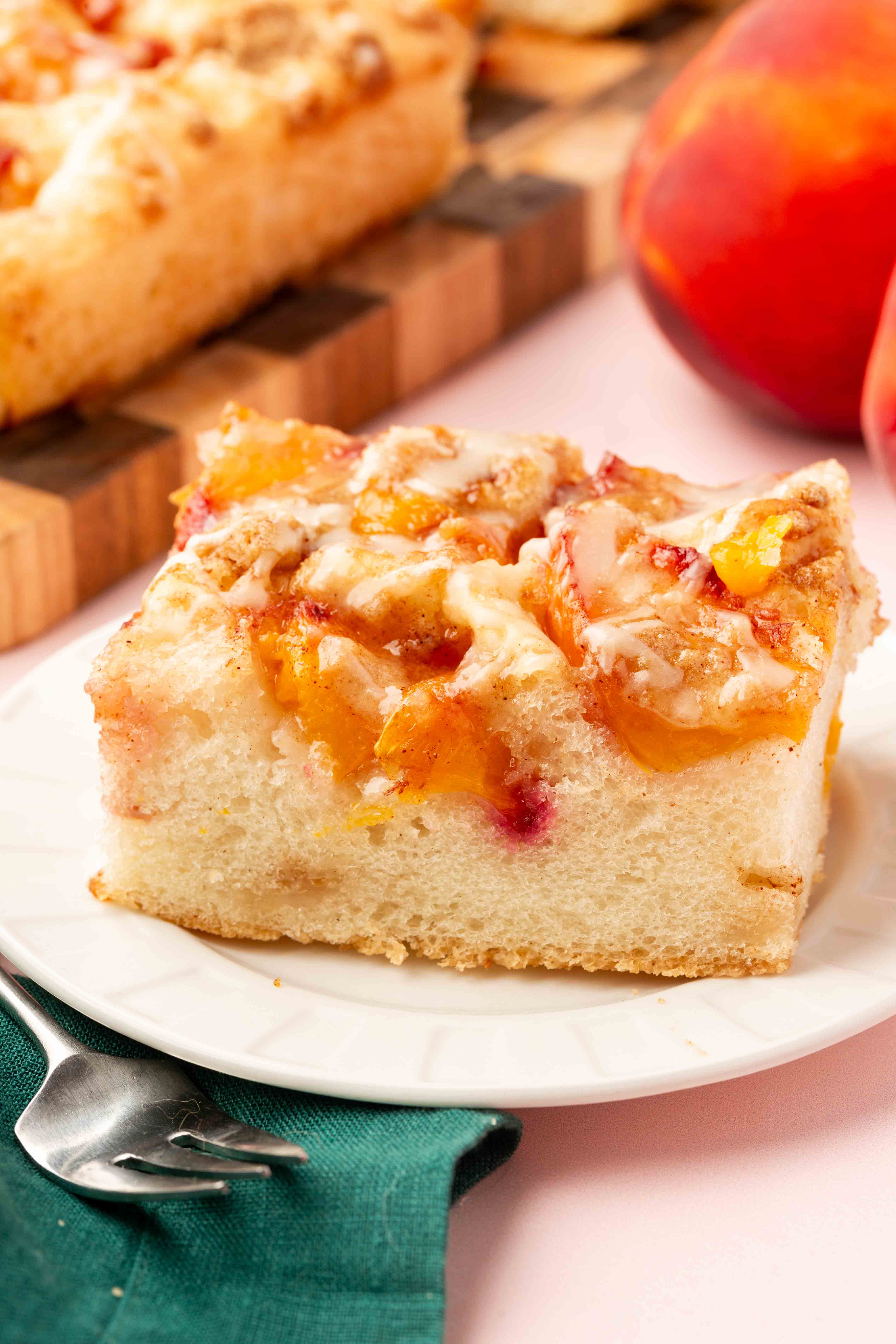 A slice of Gluten-Free Peach Crumble Focaccia with chunks of peach on top sits on a white plate next to a green napkin and fork, with a peach and more cake in the background.