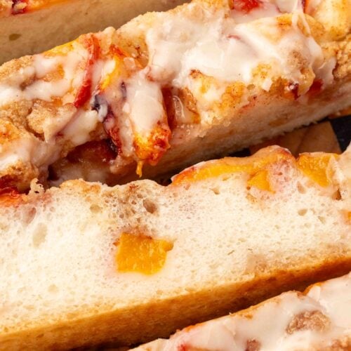 Close-up of a Gluten-Free Peach Crumble Focaccia topped with icing, crumbly streusel, and pieces of fruit—likely peach and cherry—showing a soft, airy texture inside.