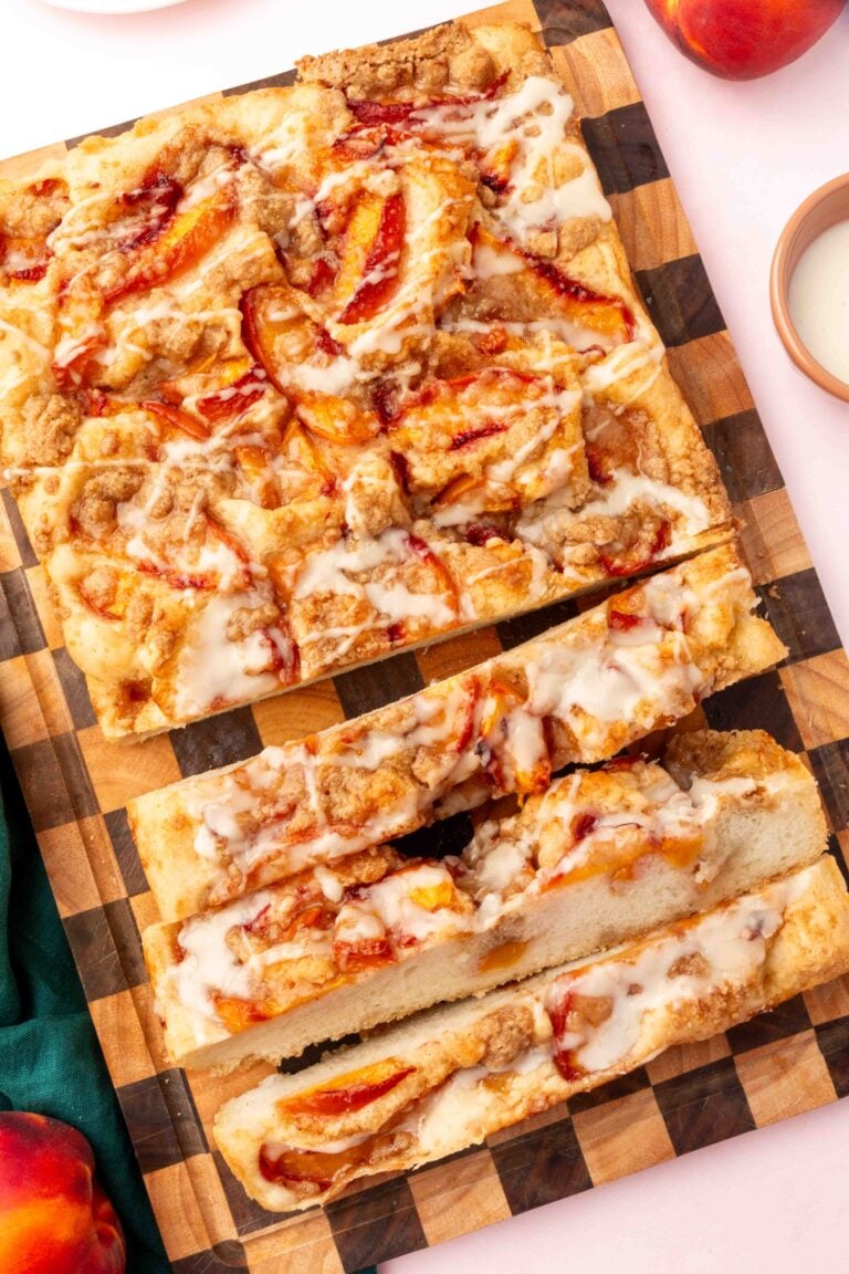 Rectangular Gluten-Free Peach Crumble Focaccia coffee cake with icing, sliced on a checkered wooden board. Cake has visible peach slices and streusel topping, surrounded by peaches and a small dish of icing.