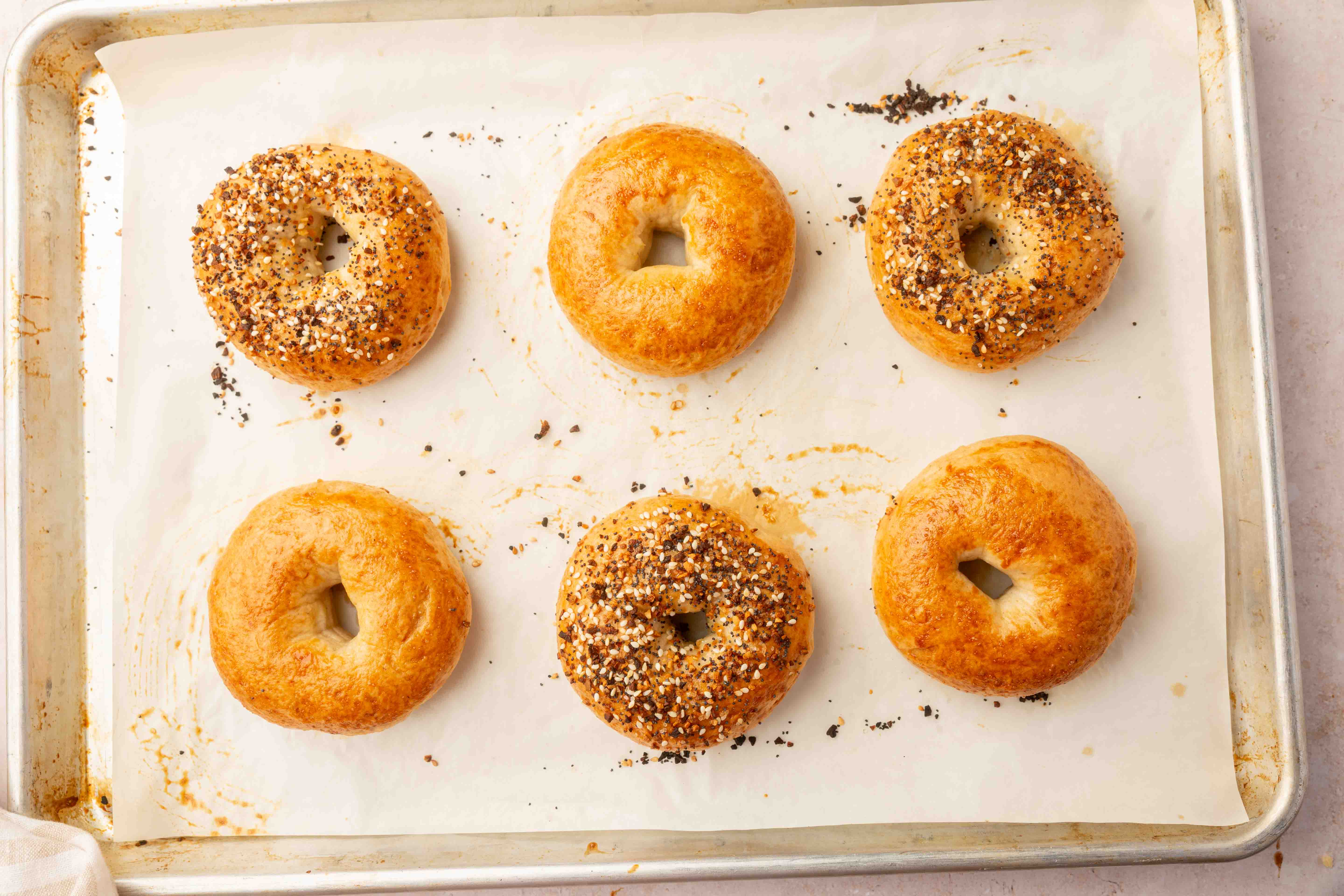 Six golden brown gluten-free bagels, three topped with everything bagel seasoning and three plain, are arranged on a parchment-lined baking sheet.