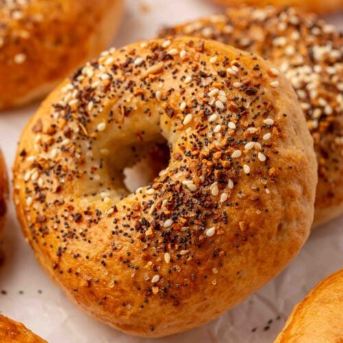 A close-up of a golden-brown gluten-free everything bagel topped with sesame seeds, poppy seeds, and seasoning, surrounded by other bagels on a white surface.