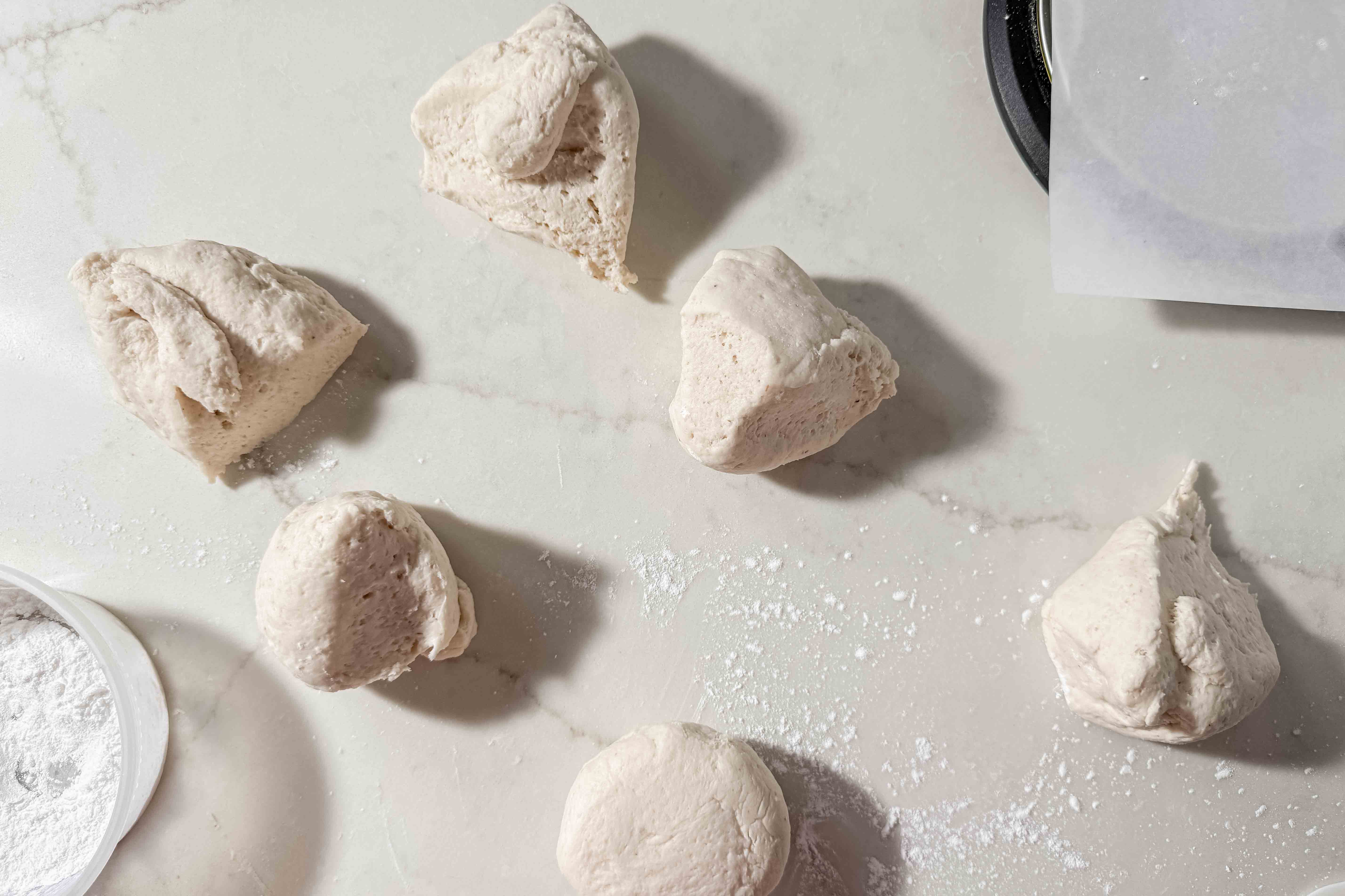 Six pieces of raw gluten-free dough are scattered on a floured white countertop, with a container of tapioca flour and a baking tray partially visible in the corners.