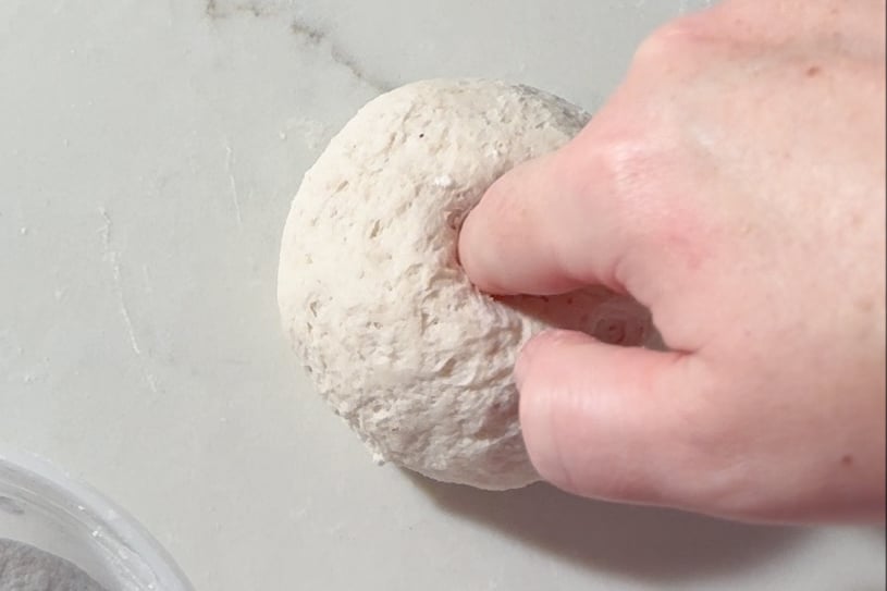 A hand presses a finger into a round ball of gluten-free bagel dough on a light-colored surface, testing its texture.