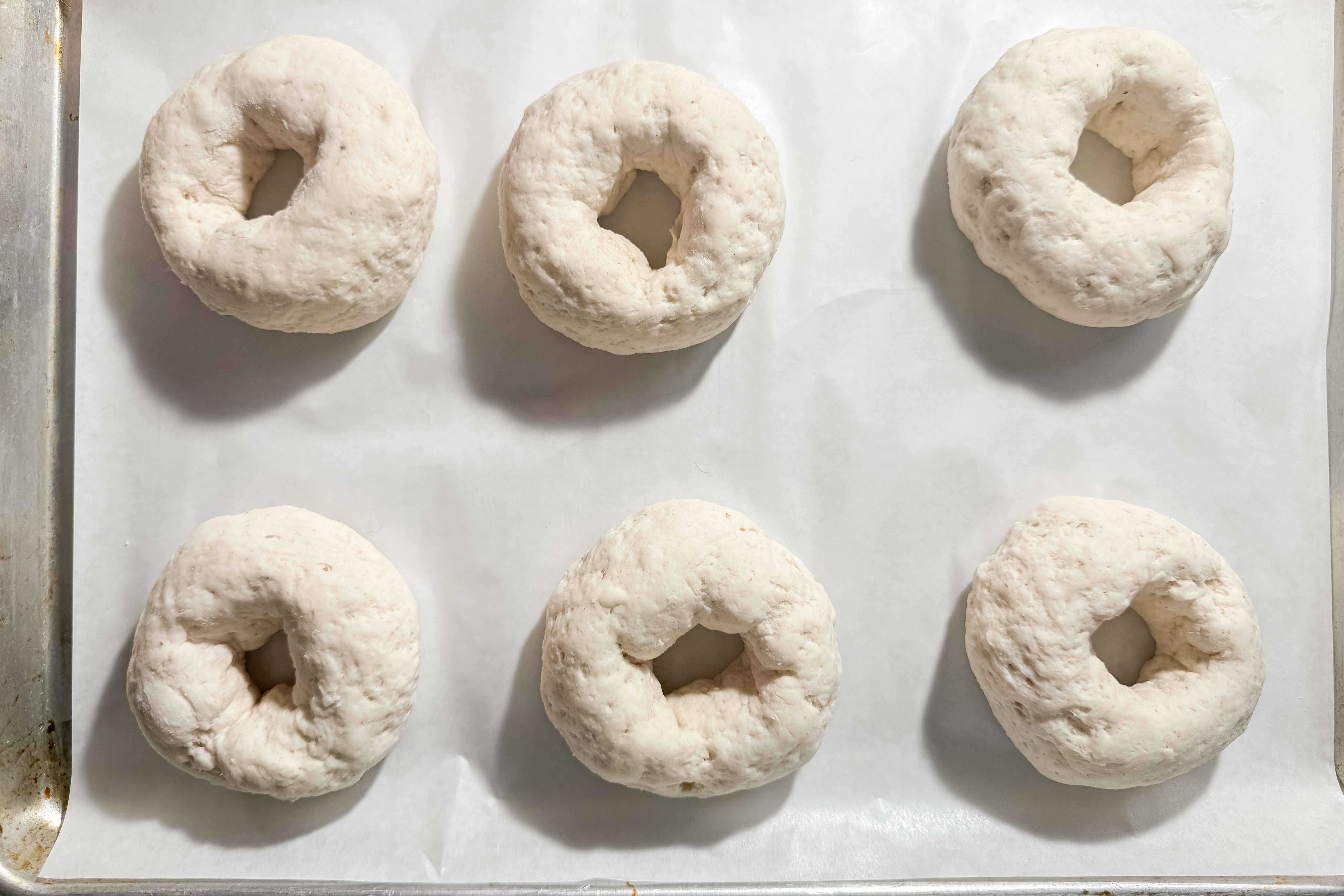 Six proofed unbaked bagels made from gluten-free dough are arranged in two rows of three on a parchment-lined baking sheet, ready for baking.