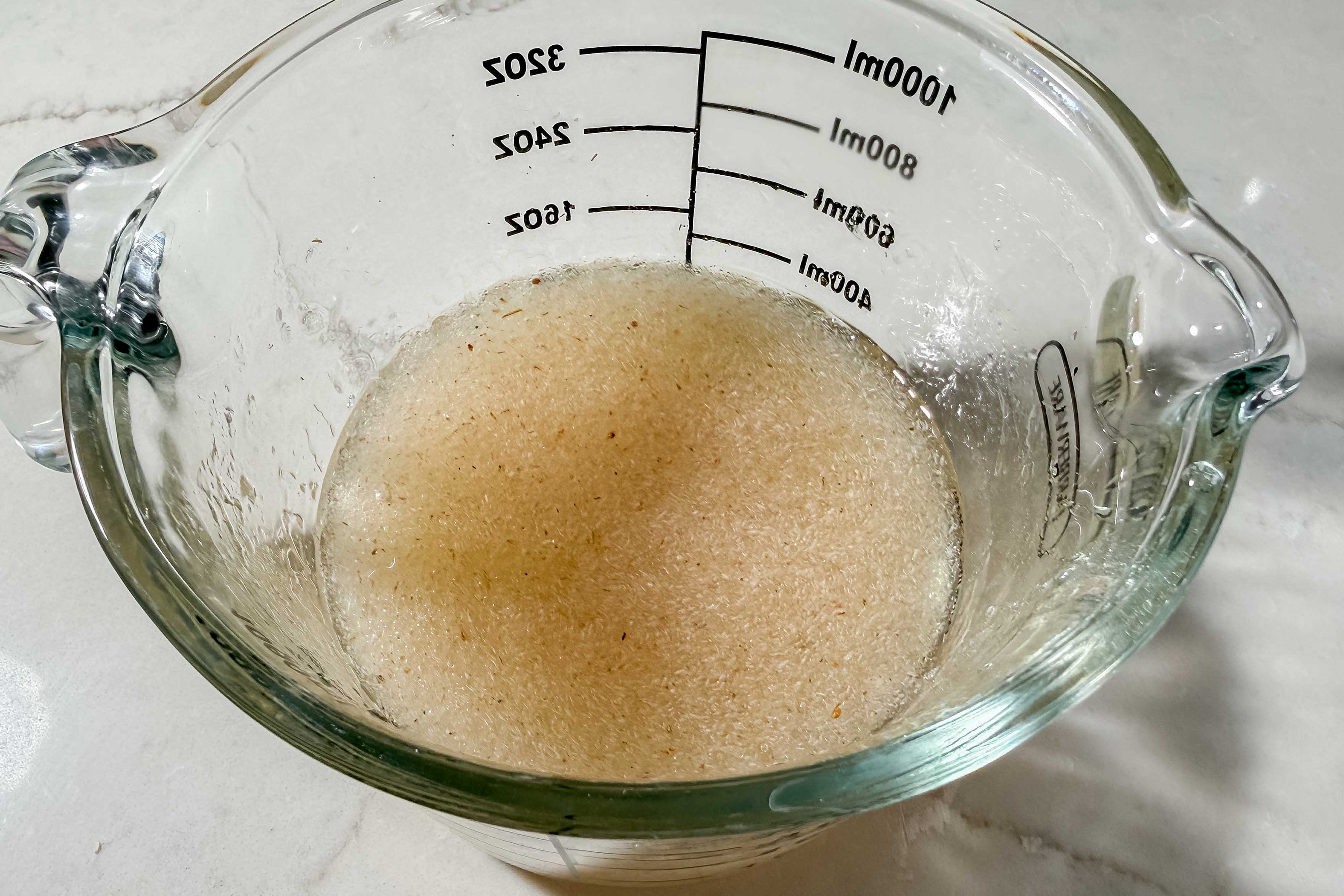A glass measuring cup containing psyllium husk mixed with water on a white countertop. Measurement markings on the side indicate volumes in milliliters and ounces.