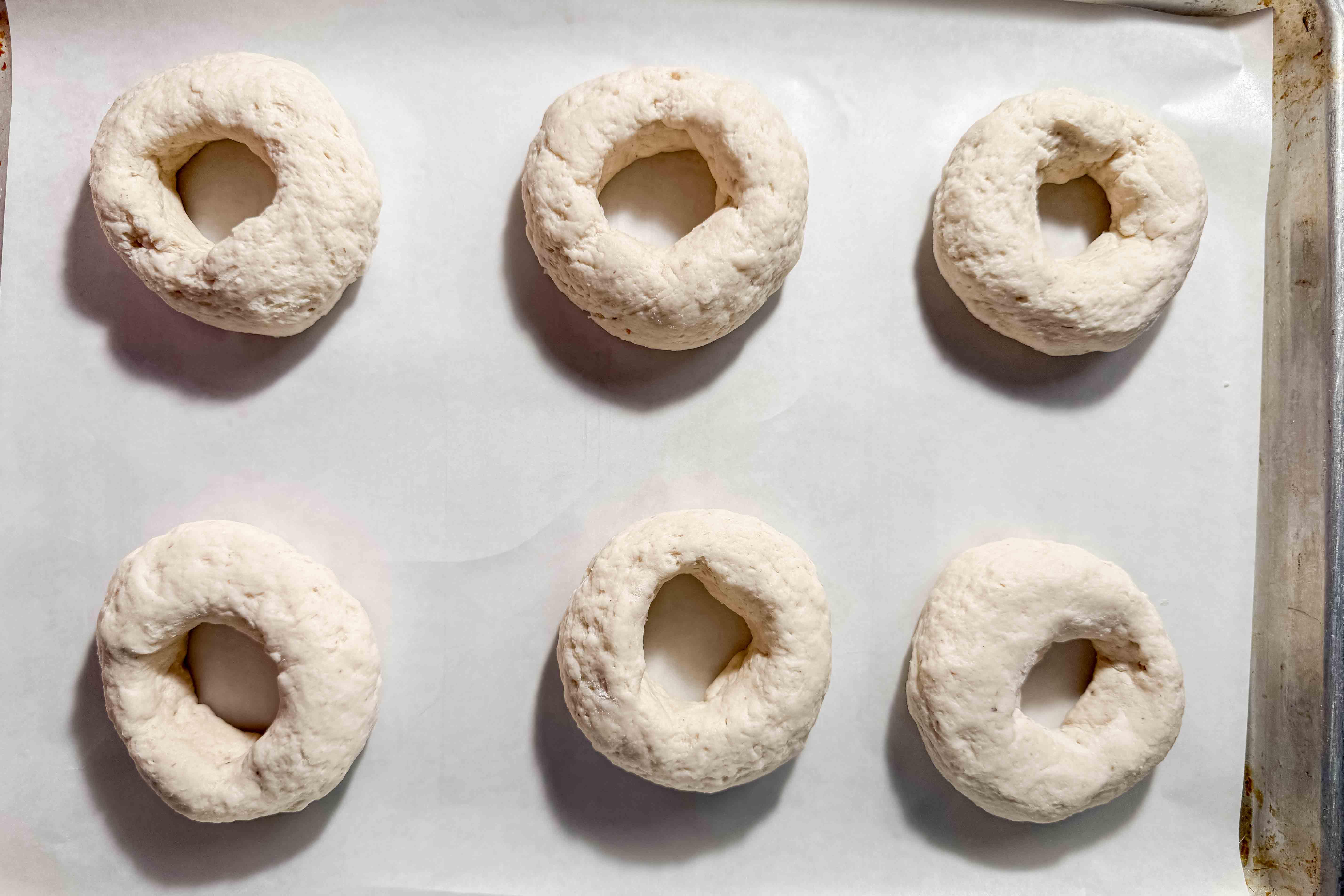 Six unproofed unbaked gluten-free bagels are arranged on a parchment-lined baking sheet. Each bagel has a hole in the middle and appears to be made from raw dough.