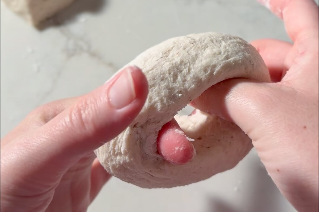 Two hands shape a piece of dough into a ring on a lightly floured surface, preparing to form a bagel.